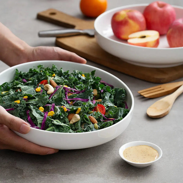 Person holding a bowl of kale salad with fruits and utensils on a table