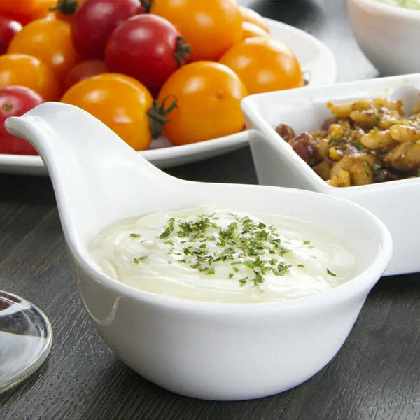 White bowl of creamy dip with herbs on a table with tomatoes and other food.