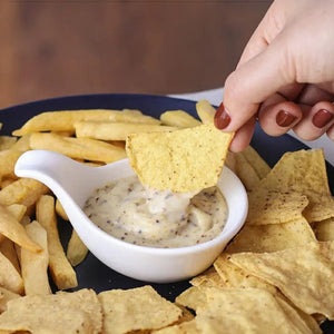 Person dipping a tortilla chip into a white bowl of dip with chips and fries on a dark surface.