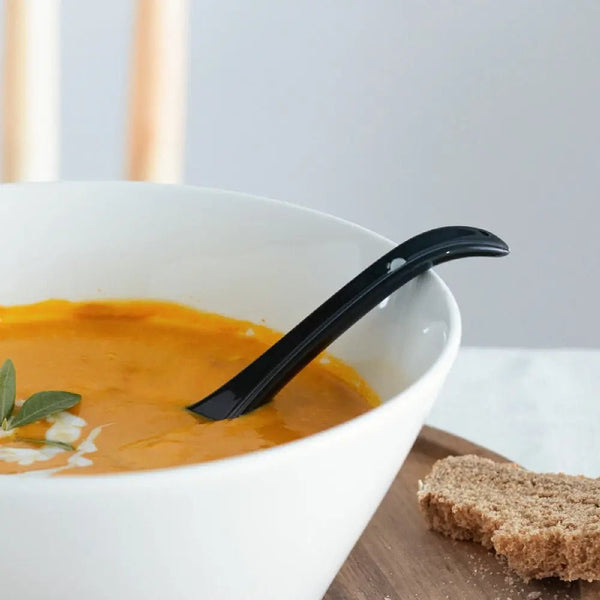 White bowl of soup with a black spoon, garnished with a sprig of sage, on a white background.