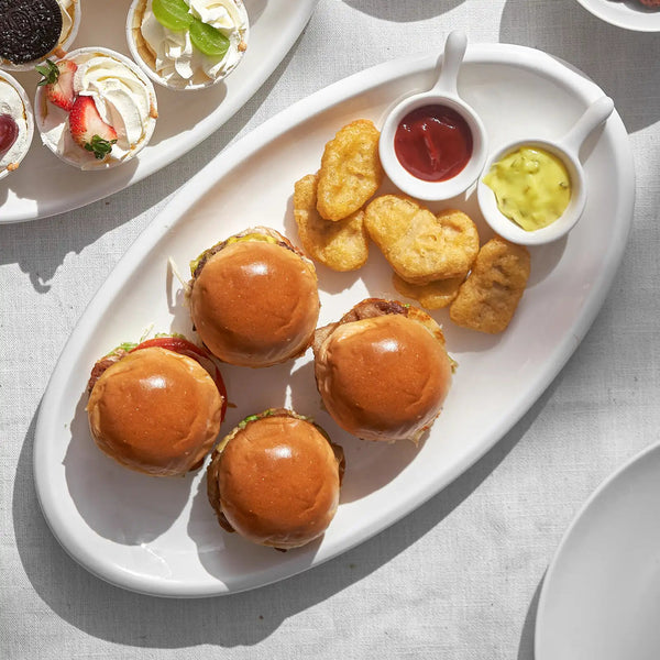 ceramic serving platter of mini burgers and tater tots with side dishes on a light gray surface