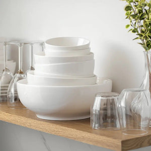 Stack of white bowls on a wooden shelf with glassware and a plant in the background.
