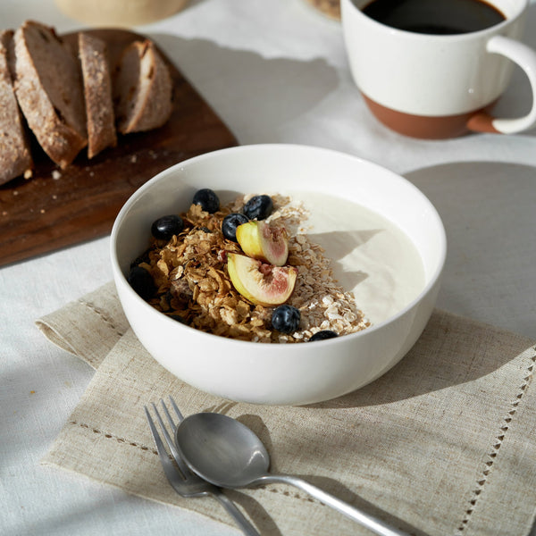 Bowl of granola with fruits on a table with bread and coffee.