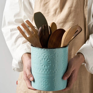 Person holding a turquoise container with wooden utensils against a neutral background