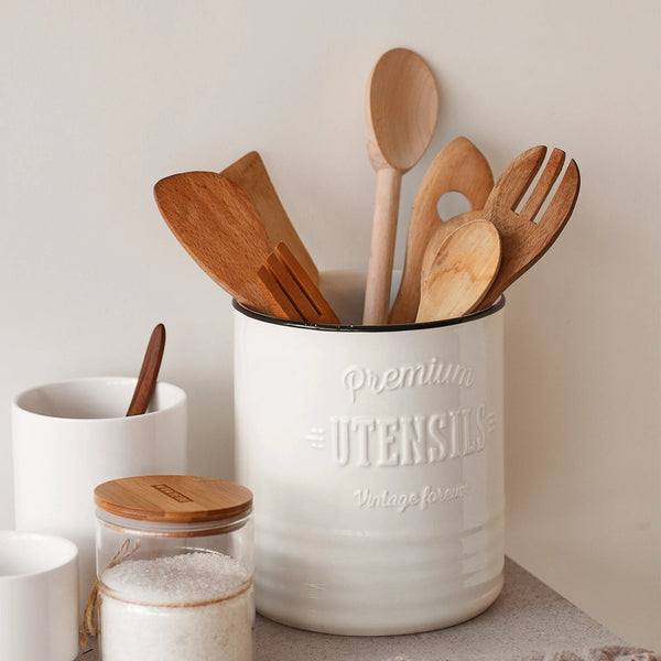 Farmhouse white utensil holder with wooden spoons and a glass container on a neutral background