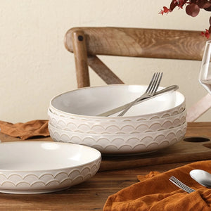 White stoneware pasta bowls with decorative patterns on a wooden table with a chair and orange napkin in the background.