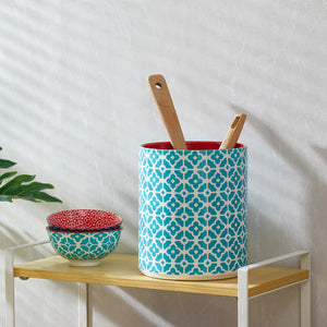 Decorative patterned ceramic utensil holder with wooden utensils on a wooden shelf against a white wall.