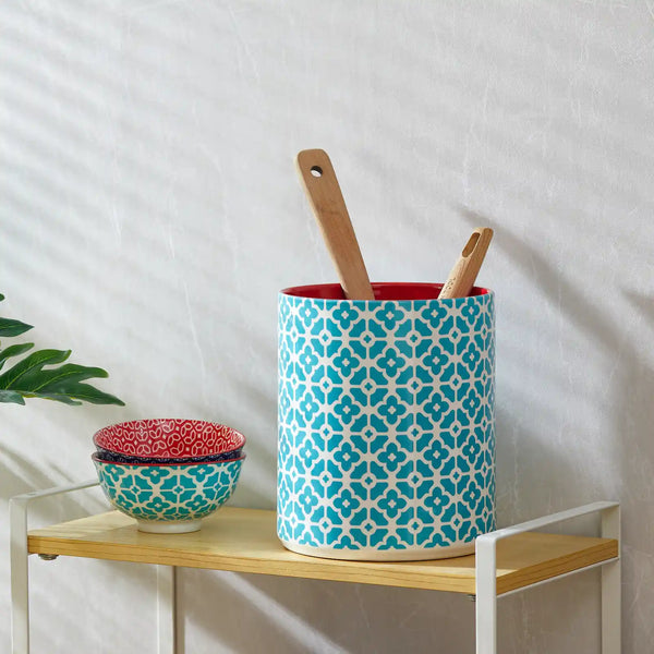 Decorative patterned ceramic utensil holder with wooden utensils on a wooden shelf against a white wall.