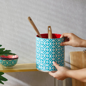Person holding a blue patterned utensil holder with red interior, wooden spoons inside, on a neutral background.