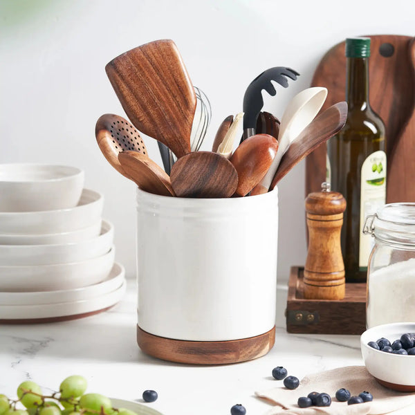 Set of wooden kitchen utensils in a white container on a kitchen counter with various items.
