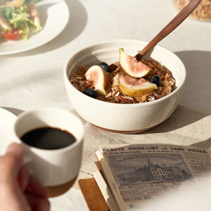 Bowl of cereal with figs and blueberries, cup of coffee, and newspaper on a table.