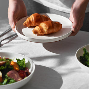 Person holding a plate with two croissants on a light surface with bowls of salad in the background.