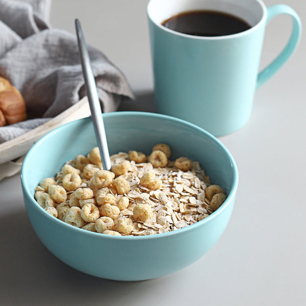 Teal bowl of cereal with a spoon and teal mug of coffee on a light surface.