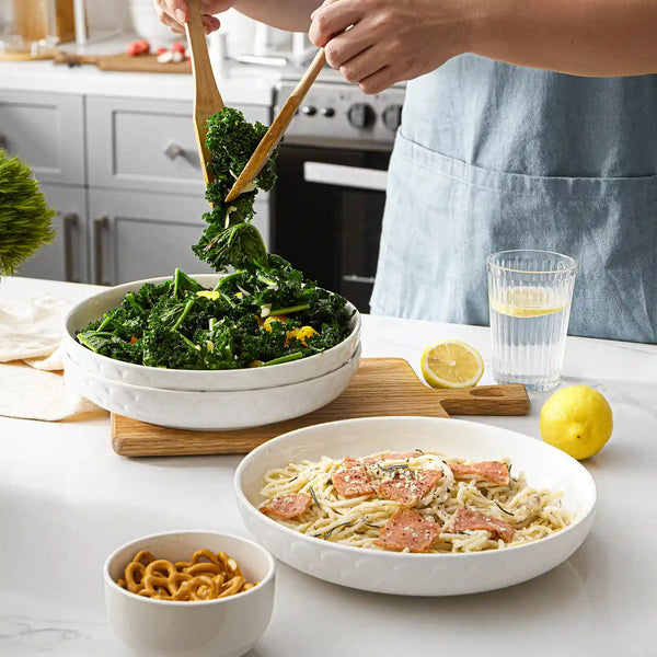Person serving food from a white bowl of greens onto a plate of pasta with salmon on a kitchen counter.