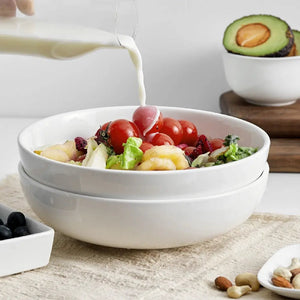 White bowl with salad being poured into it, surrounded by avocados and nuts on a light surface.