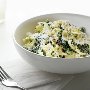 White bowl of pasta with greens and parmesan cheese on a white surface