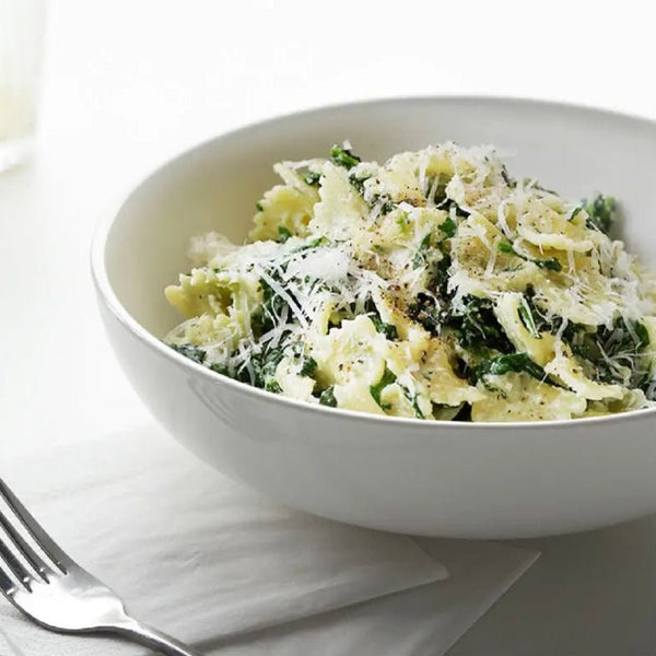 White bowl of pasta with greens and parmesan cheese on a white surface