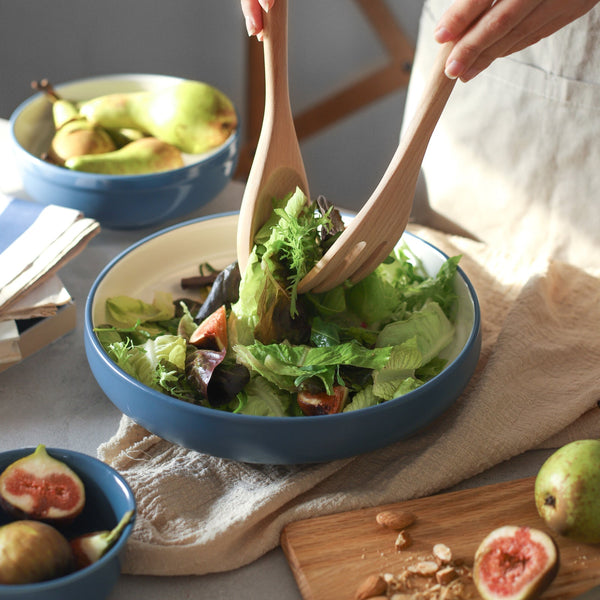 Person tossing a salad in a blue bowl with wooden utensils, surrounded by fruits and a book.