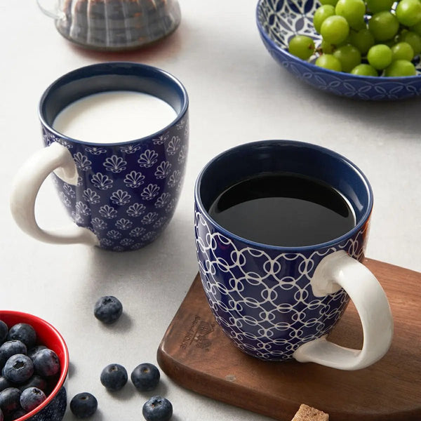 Two blue patterned mugs with one containing milk and the other coffee, on a table with fruit bowls.