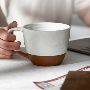 Person holding a ceramic mug with a brown bottom and white top on a table.