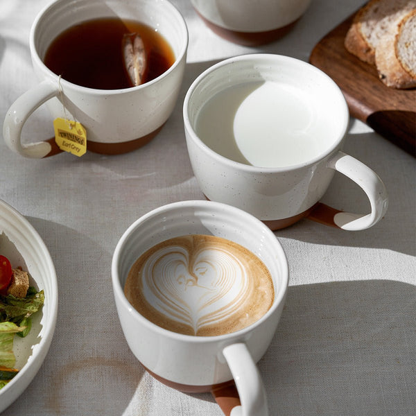 large ceramic coffee cups containing coffee, tea, and a latte art design on a light gray surface.