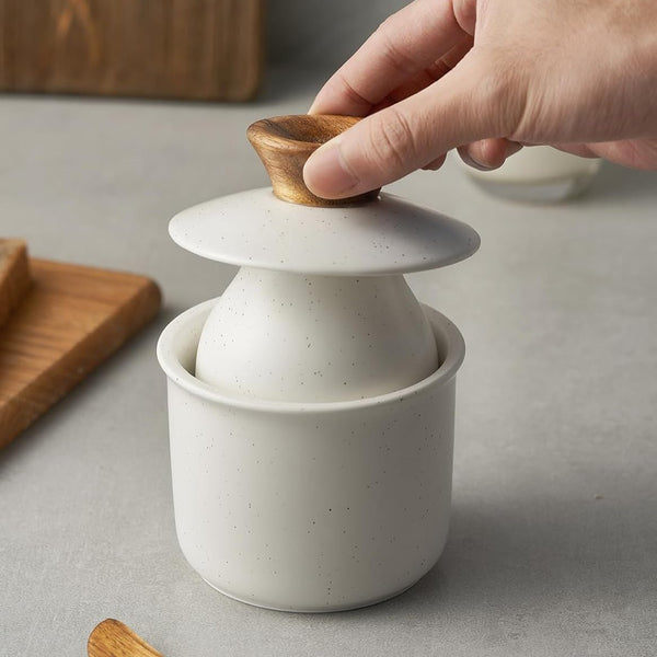 White ceramic container with wooden lid being held by a hand on a gray surface.