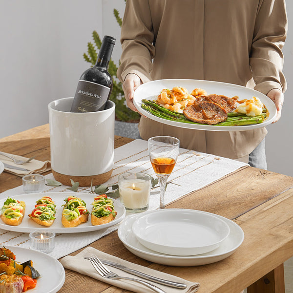 Person holding a oval serving platter of food with a bottle of wine on a table set for dinner.