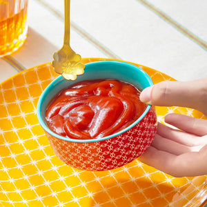Hand dipping a golden spoon into a bowl of ketchup on a yellow checkered tablecloth.