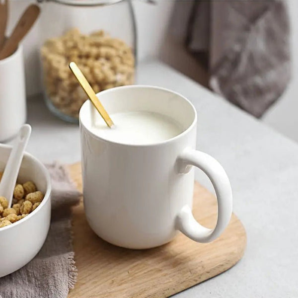 A white mug with milk and a spoon inside, placed on a wooden coaster, surrounded by cereal and jars.