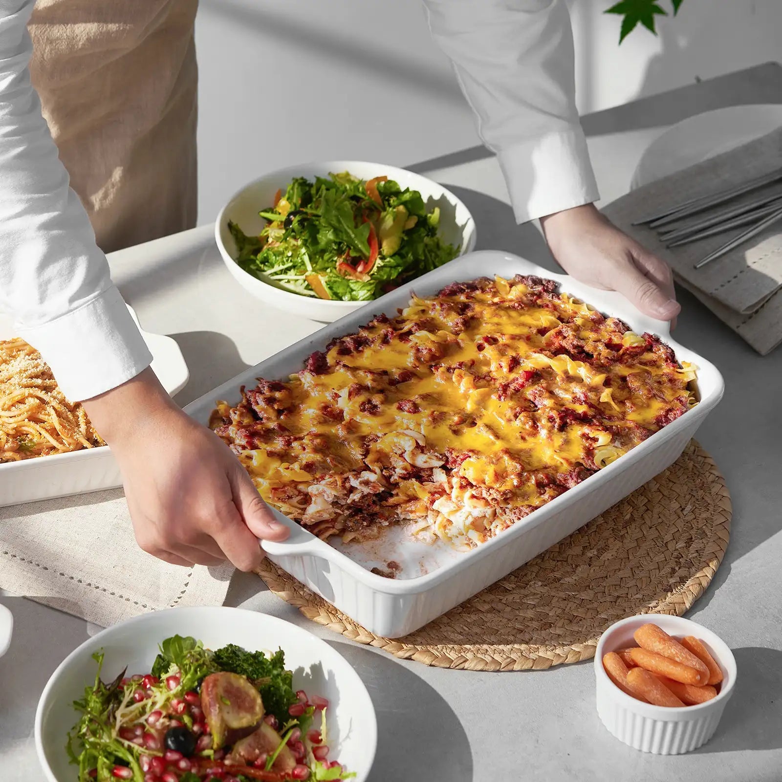 Person serving a dish of cheesy casserole with salads and carrots on a table.