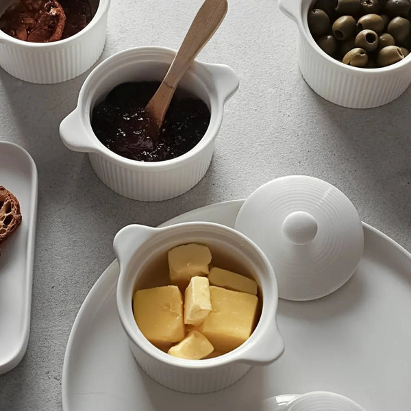 White ceramic ramekins containing butter and a dark spread, with a wooden spoon, on a light gray surface.