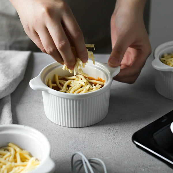 Person adding cheese  to a small white ramekin on a gray surface.