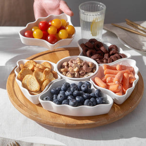 Assorted snacks including fruits, nuts, and bread on a wooden tray with a glass of water in the background.