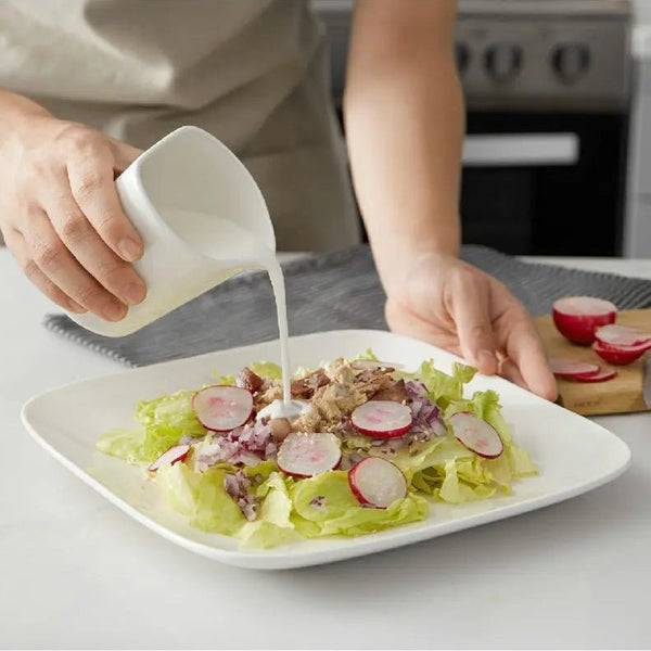 Person pouring dressing onto a salad on a white plate in a kitchen setting.