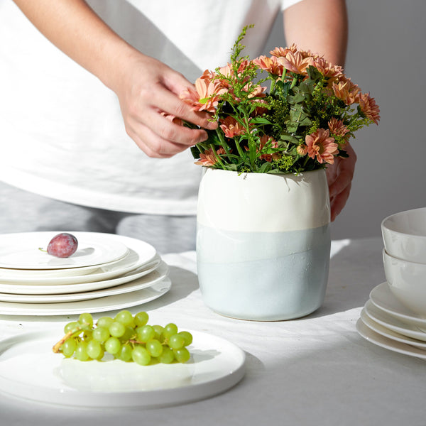 Person arranging flowers in a blue and white utensil crock on a table with plates and grapes.