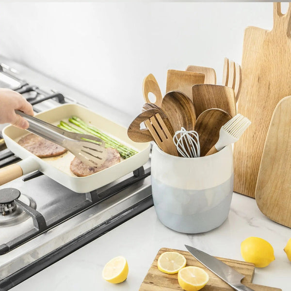 Kitchen scene with cooking utensils in a container, lemons on a cutting board, and food being prepared.