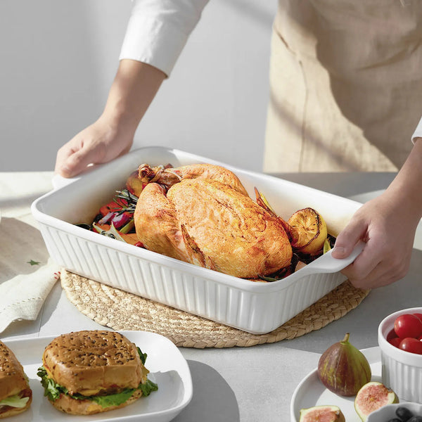 Person holding a white ceramic roasting dish with roasted chicken and vegetables on a table.