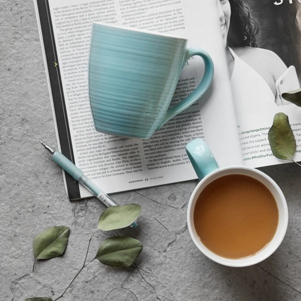 Turquoise mug with coffee on an open book with a pen and green leaves.