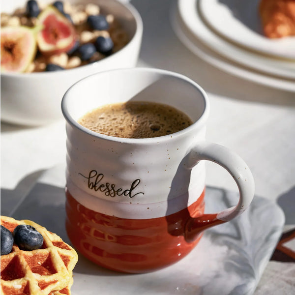 Mug with 'blessed' text filled with coffee, surrounded by a waffle and fruit on a table.