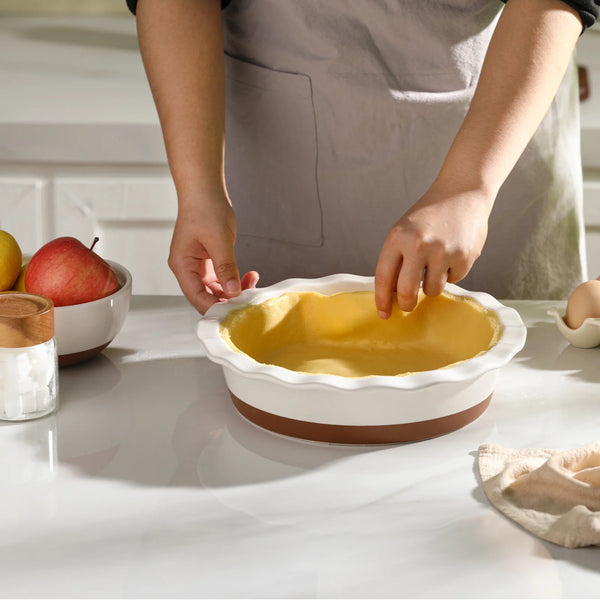 Person preparing a pie crust in a baking dish with apples and a bowl on the counter.