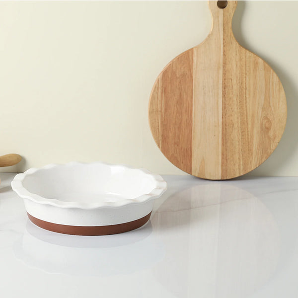 White ceramic baking dish with brown base and wooden cutting board on a light surface