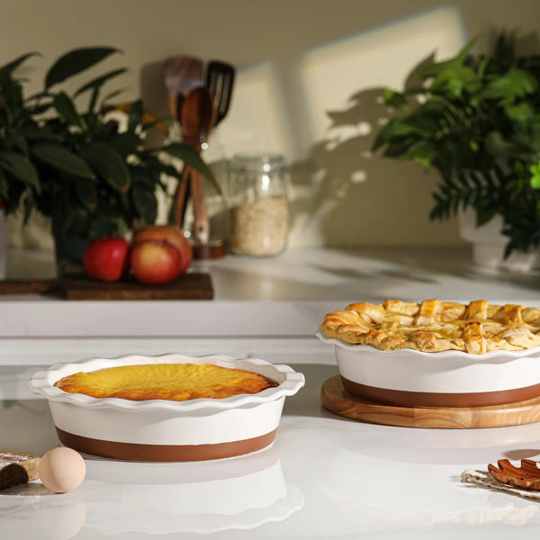Two pies in ceramic baking dishes on a kitchen counter with plants and apples in the background.