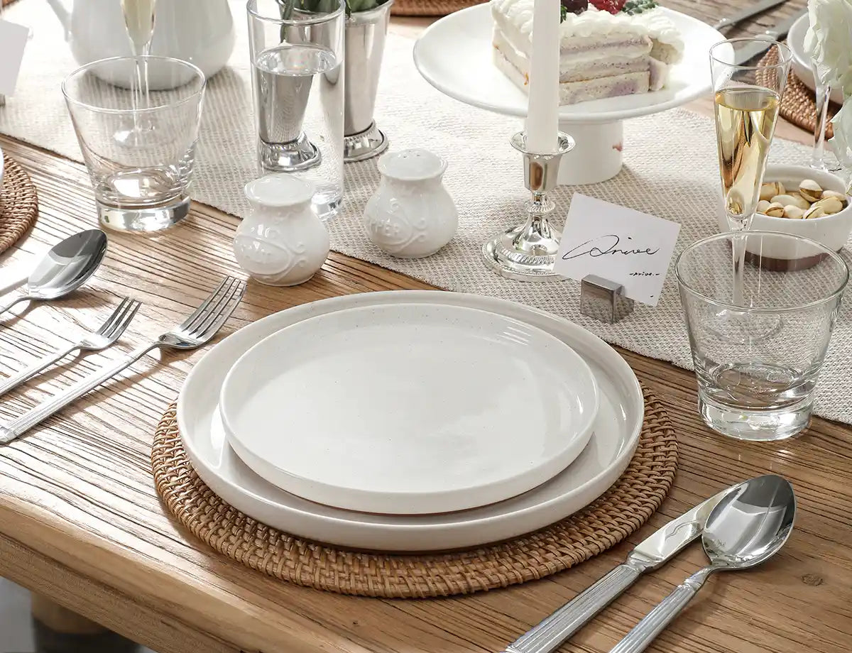 Dining table setting with white dinner plate sets, silverware, and glasses on a wooden table.