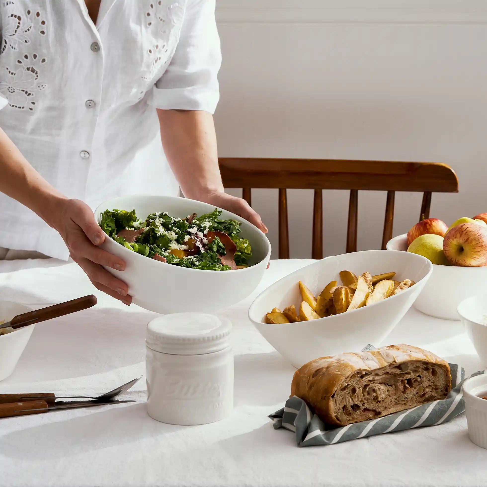 Person holding a bowl of salad at a table with bread, apples, and white French Butter Crock.