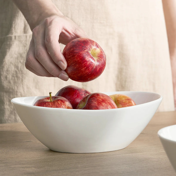 Hand placing a red apple into a white bowl on a wooden surface.