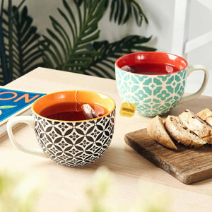 Two patterned coffee mugs with tea and breakfast on a table with a blurred background