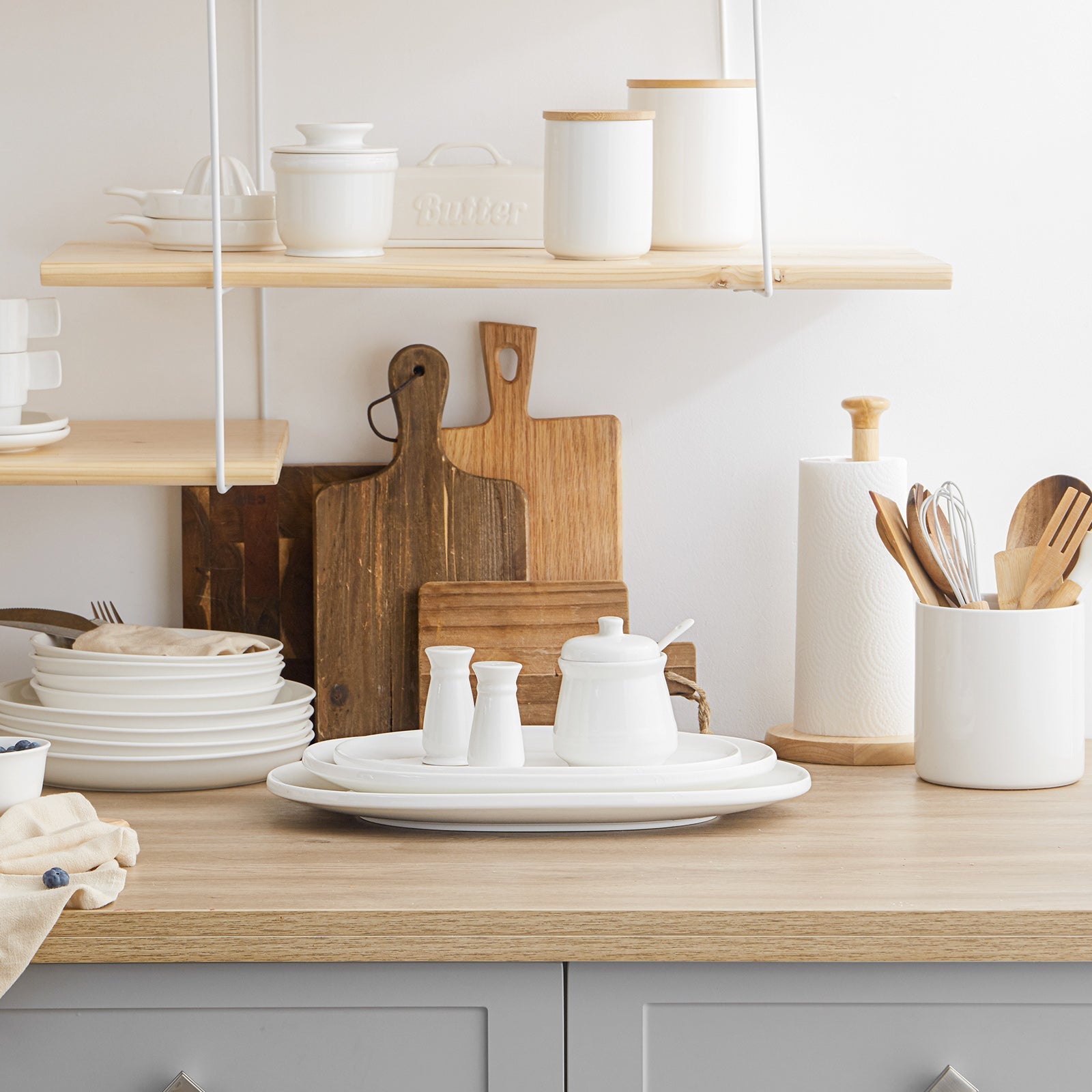 Kitchen counter with white dishes, wooden cutting boards, and kitchen utensils on a light wood surface.